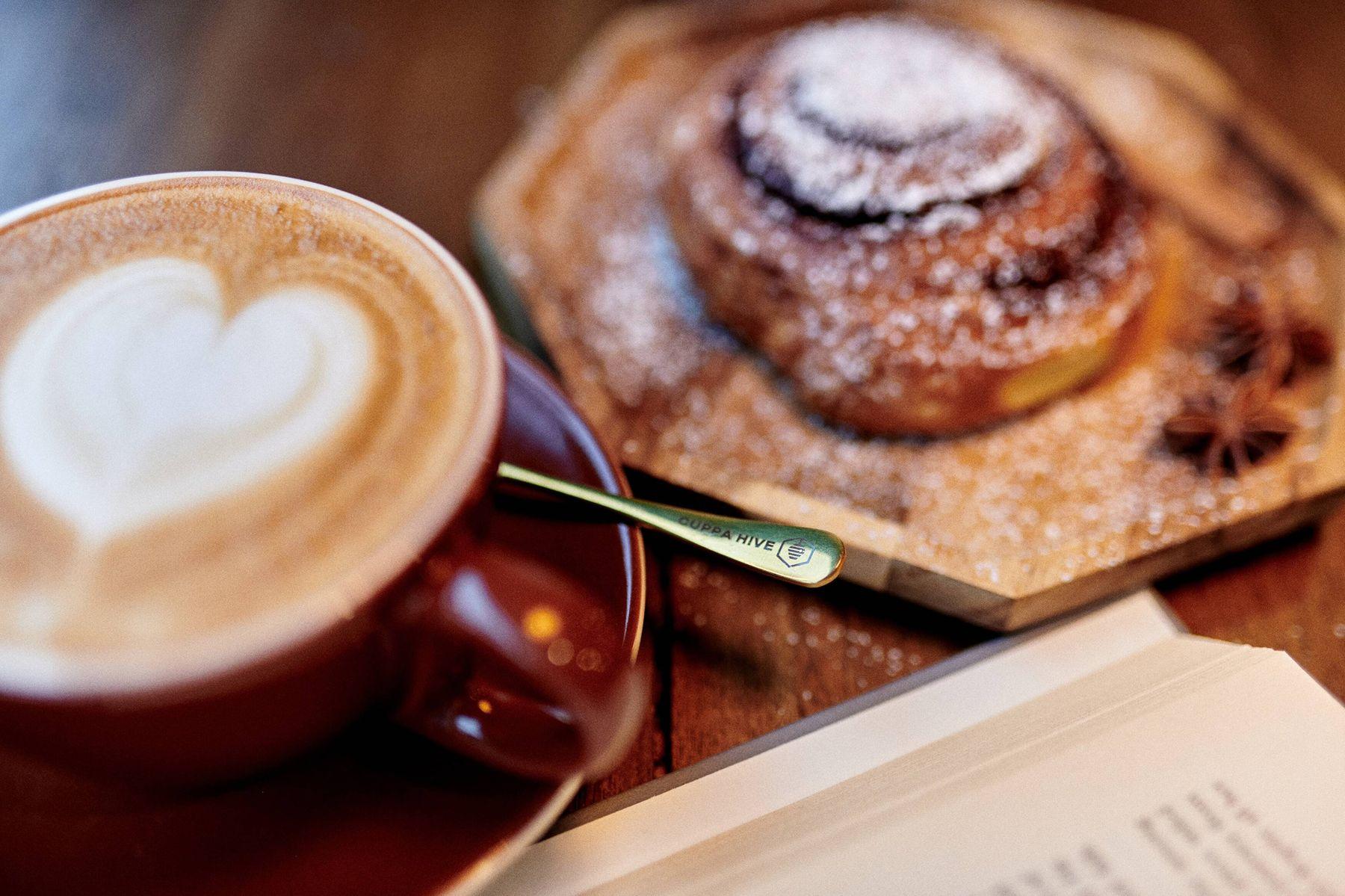 A heart-poured latte and a sugared cinnamon roll on a wood table, with an open book